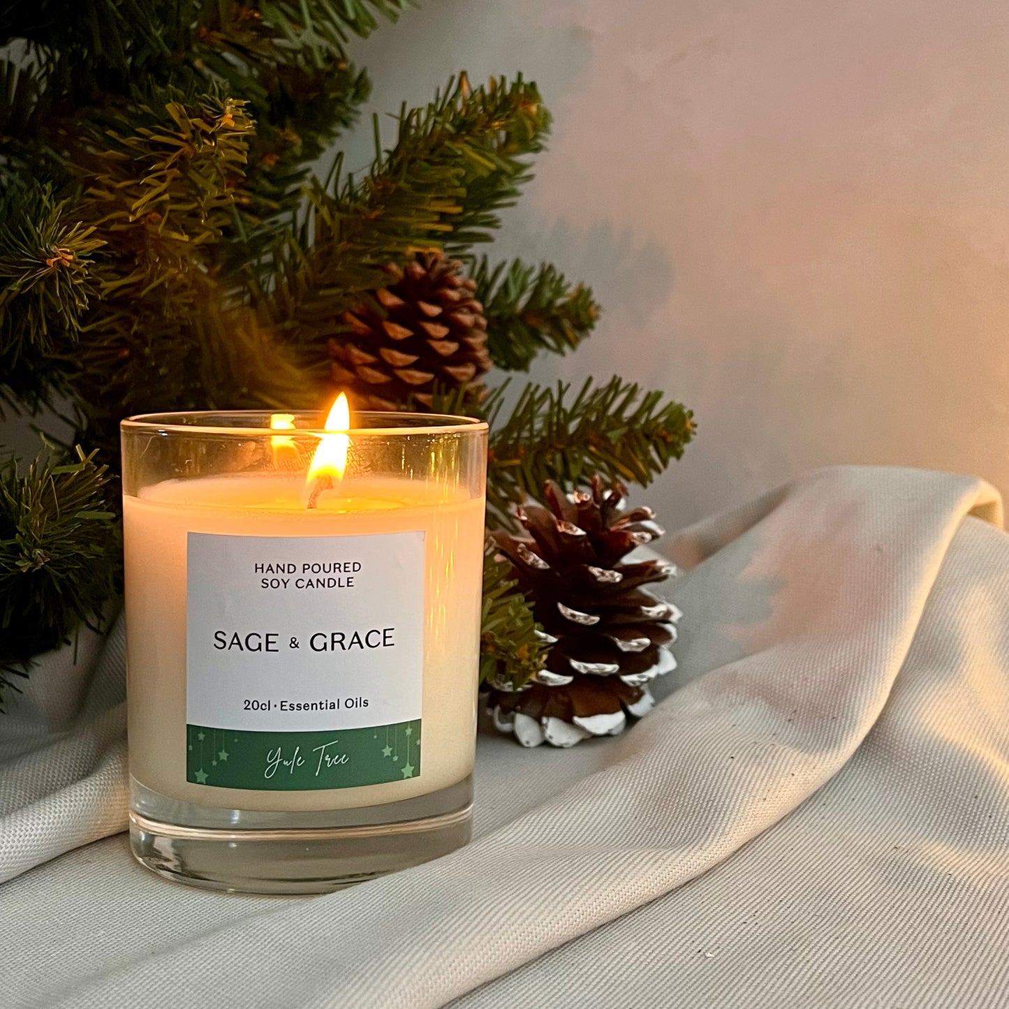 Scented candle labeled 'Sage & Grace' with a Christmas tree and pinecones in the background