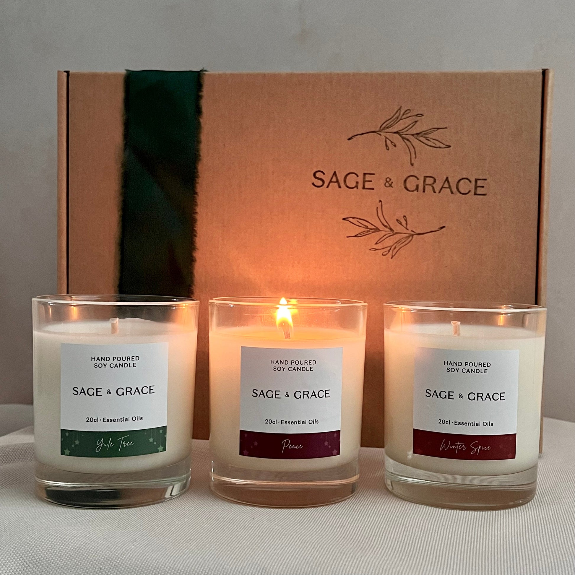 Three lit candles from Sage & Grace brand in front of a branded box on a gray surface.