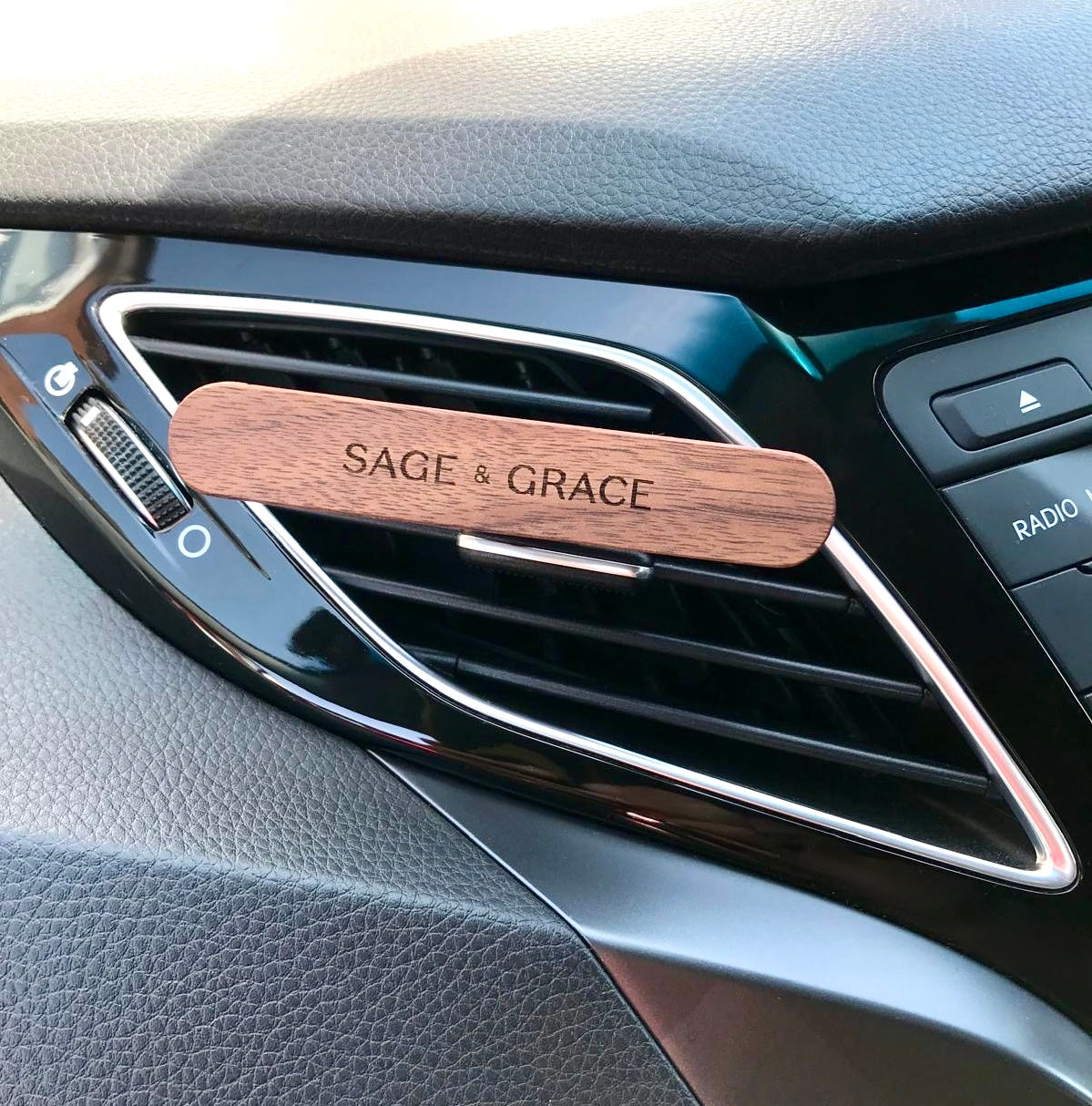 A stylish wooden car freshener, with Sage & Grace logo on it, clipped onto a car air vent